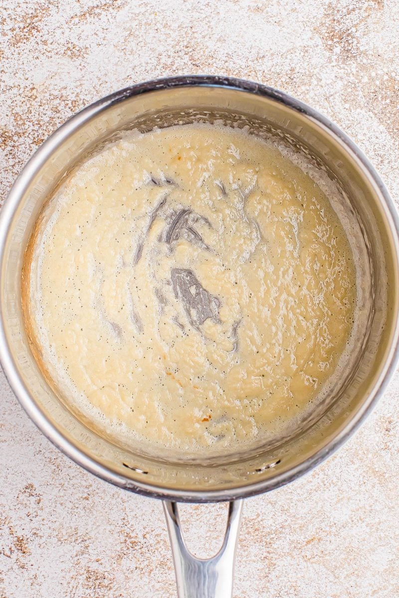 A stainless steel pot containing a thick, beige roux mixture with visible black specks, resting on a light textured surface.