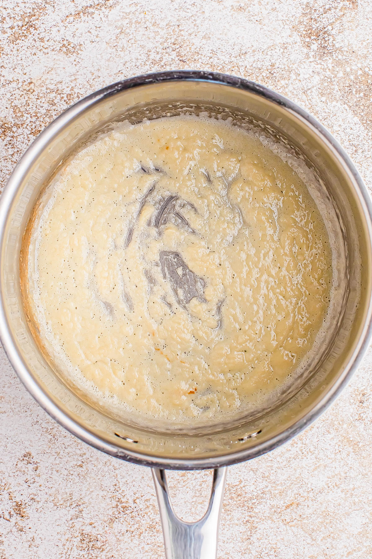 A stainless steel pot containing a thick, beige roux mixture with visible black specks, resting on a light textured surface.