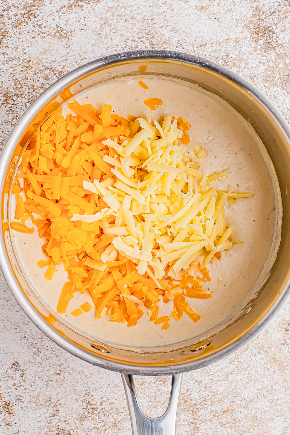 Shredded cheddar and white cheese sitting on top of a creamy sauce in a stainless steel saucepan.