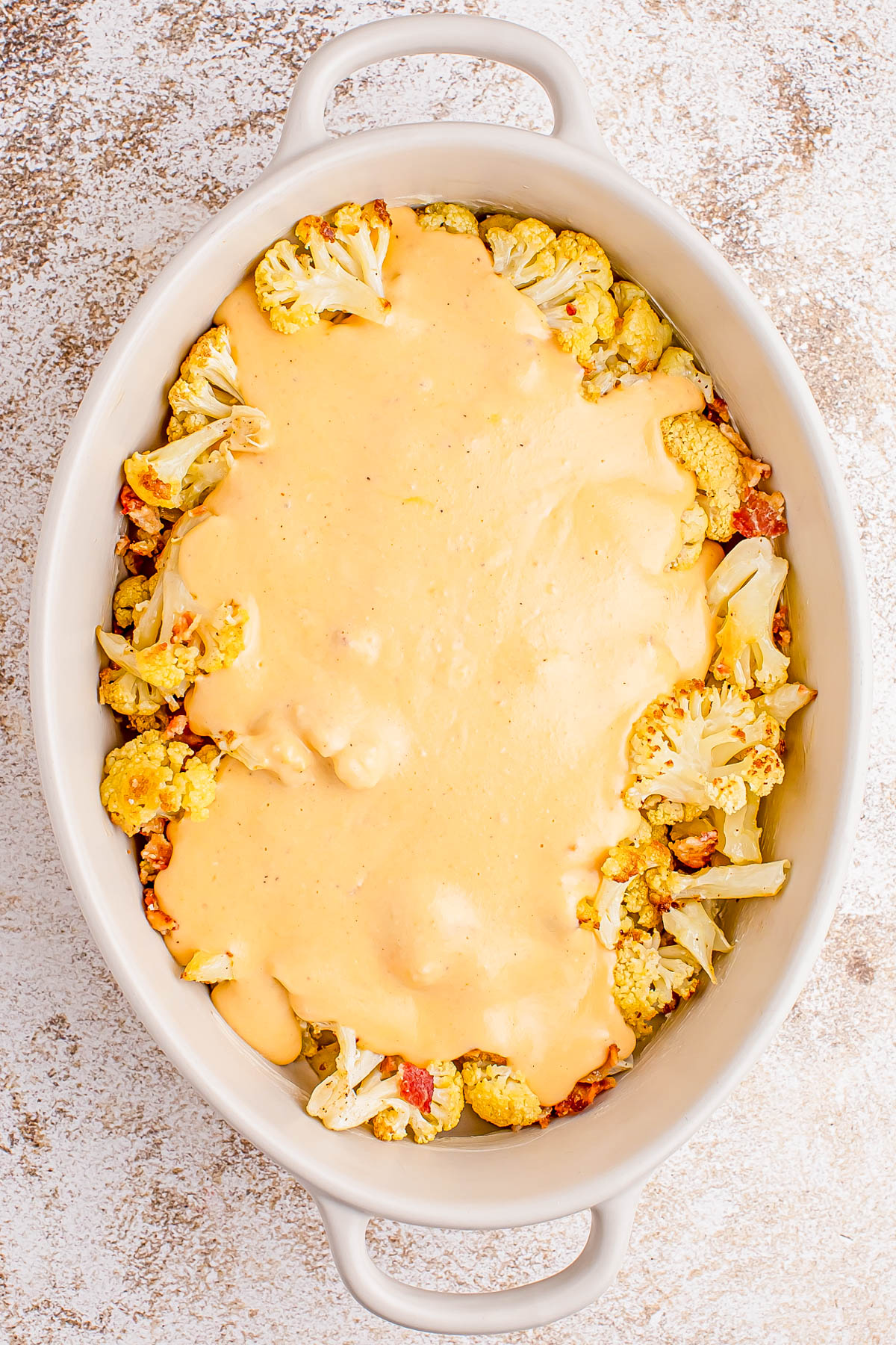 Oval baking dish filled with roasted cauliflower florets topped with a layer of melted cheese sauce, placed on a textured surface.