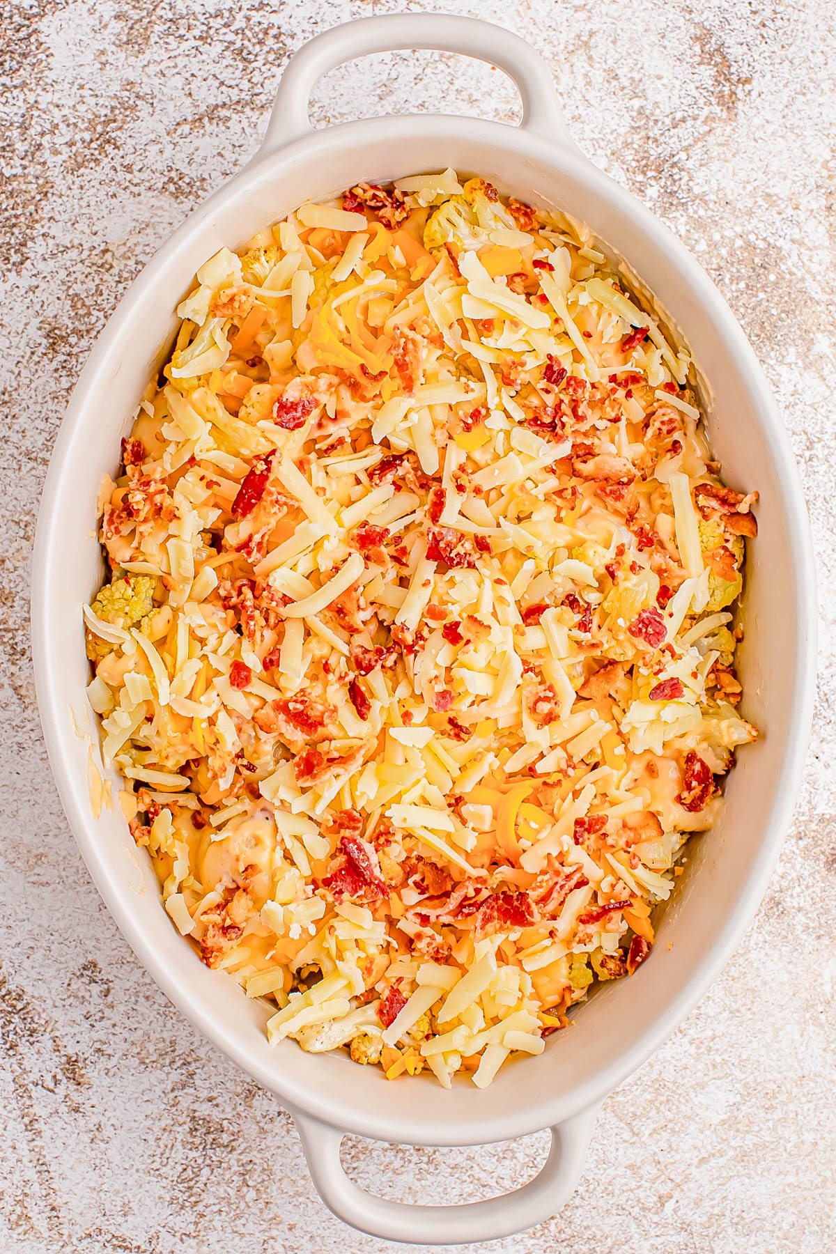 An oval baking dish filled with a mixture of shredded cheese, chopped bacon, and pasta, ready to be baked.