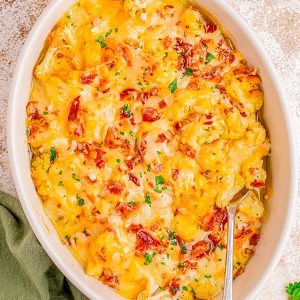 A casserole dish filled with cheesy potatoes topped with bacon bits and chopped parsley, with a serving spoon inside.