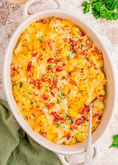 A casserole dish filled with cheesy potatoes topped with bacon bits and chopped parsley, with a serving spoon inside.