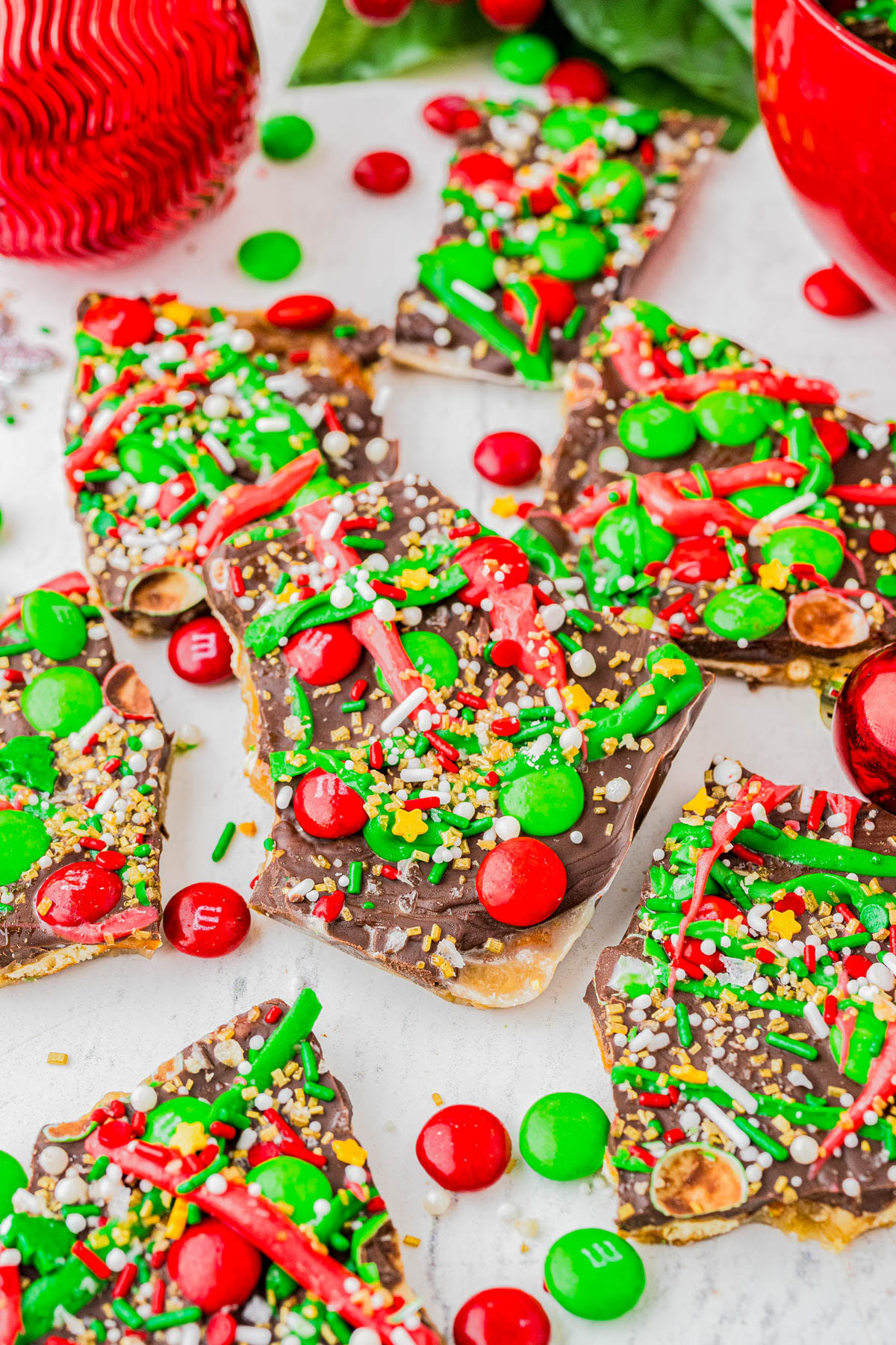 Chocolate bark topped with festive red and green candies, sprinkles, and nuts, broken into pieces and arranged on a white surface with Christmas decorations nearby.