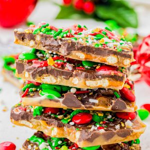 A stack of holiday toffee bark pieces topped with chocolate, red and green candy-coated chocolates, pretzel bits, and festive sprinkles sits on a white surface.
