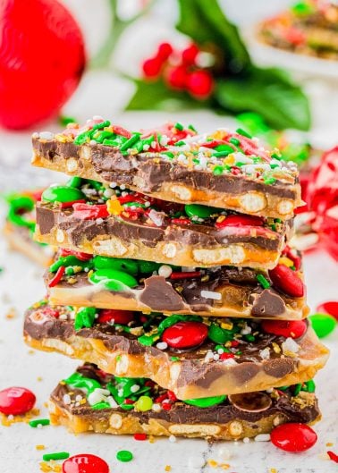 A stack of holiday toffee bark pieces topped with chocolate, red and green candy-coated chocolates, pretzel bits, and festive sprinkles sits on a white surface.