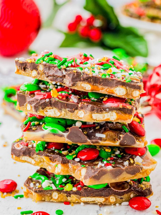 A stack of holiday toffee bark pieces topped with chocolate, red and green candy-coated chocolates, pretzel bits, and festive sprinkles sits on a white surface.