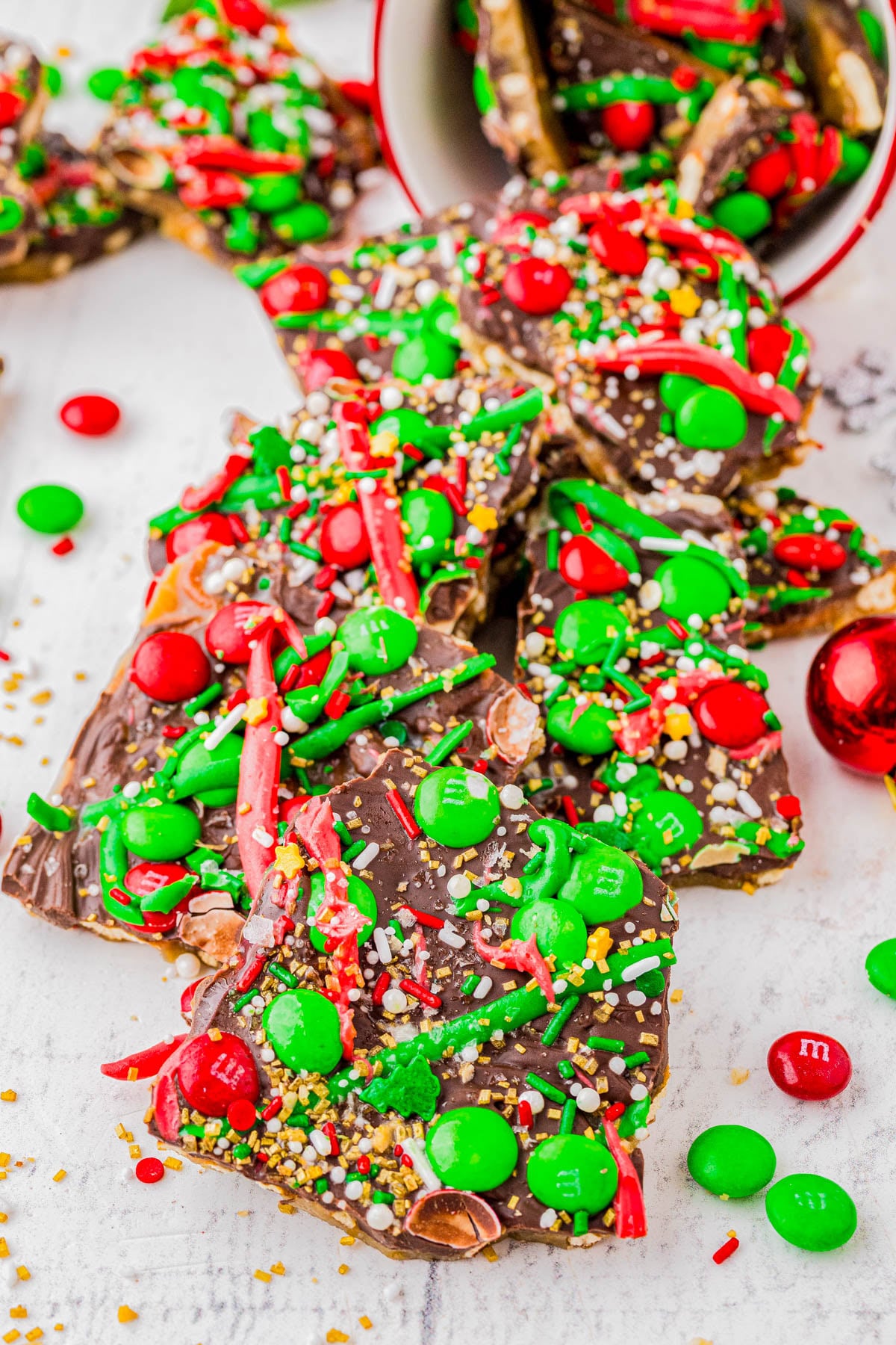 Chocolate bark topped with red and green M&Ms, sprinkles, and drizzle, displayed on a white surface with some candies scattered around.