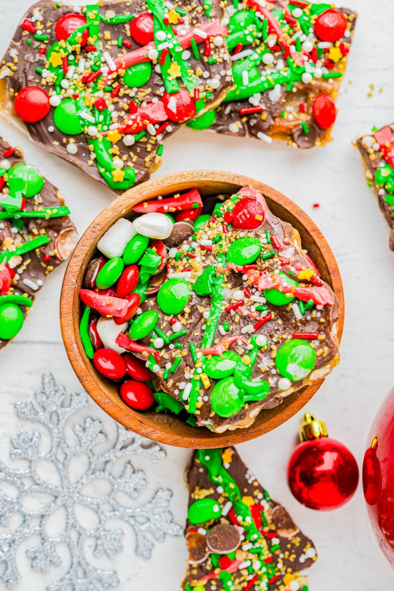 A bowl and pieces of festive holiday bark topped with green, red, and white candy, sprinkles, and chocolate, with Christmas ornaments nearby.