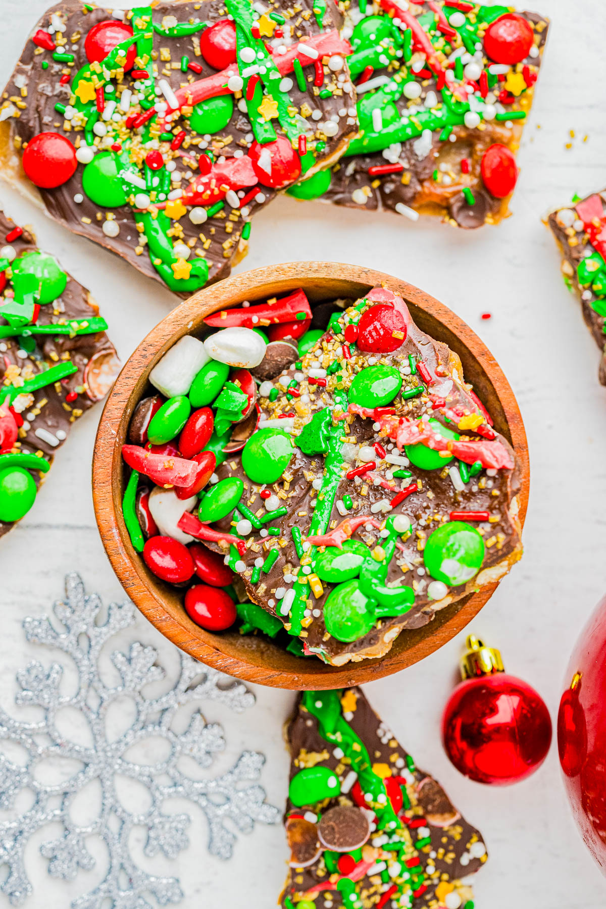 A bowl and pieces of festive holiday bark topped with green, red, and white candy, sprinkles, and chocolate, with Christmas ornaments nearby.