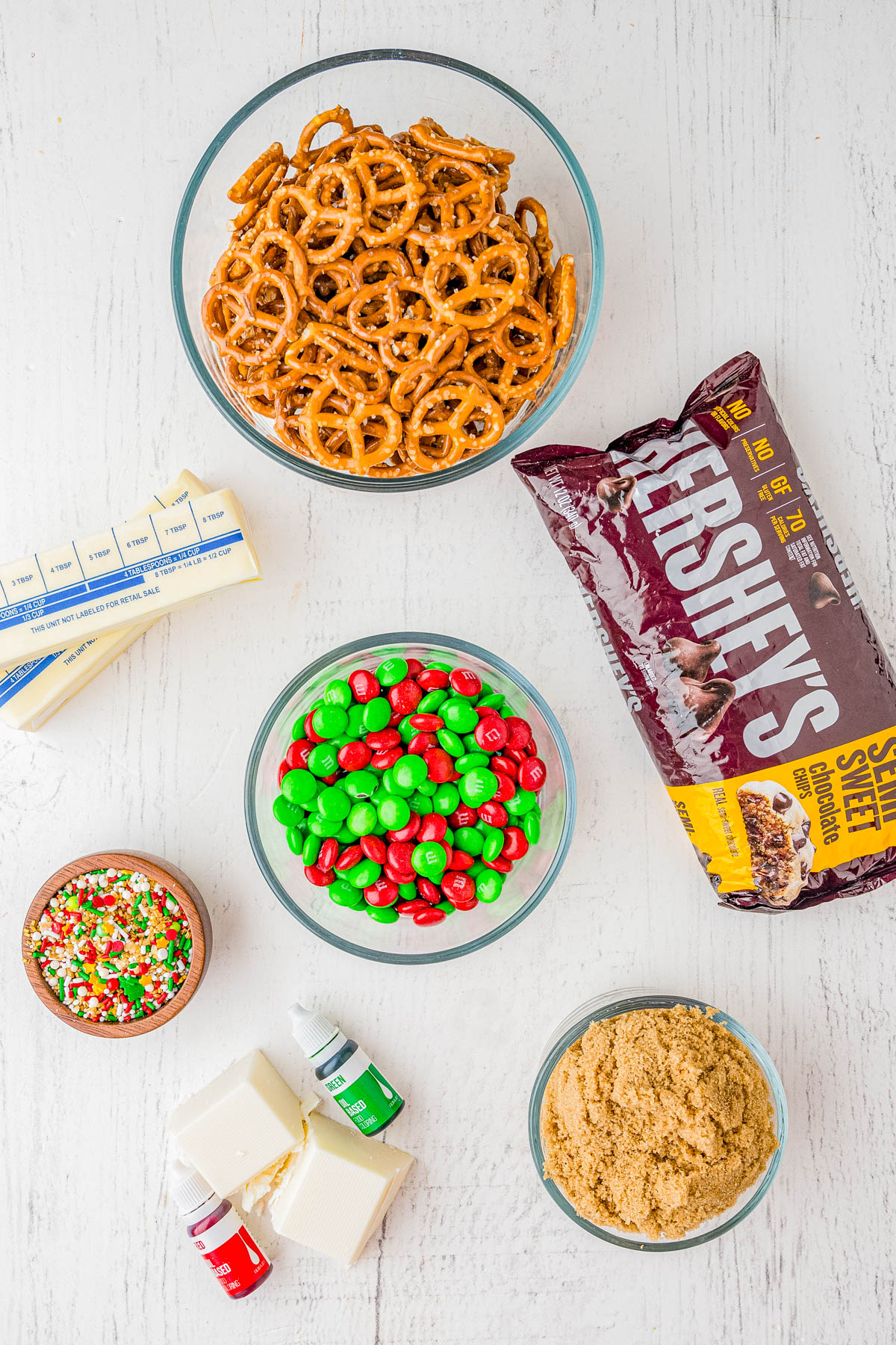 Assorted baking ingredients on a white surface, including pretzels, M&M's, chocolate chips, brown sugar, sprinkles, butter, almond bark, and food coloring.