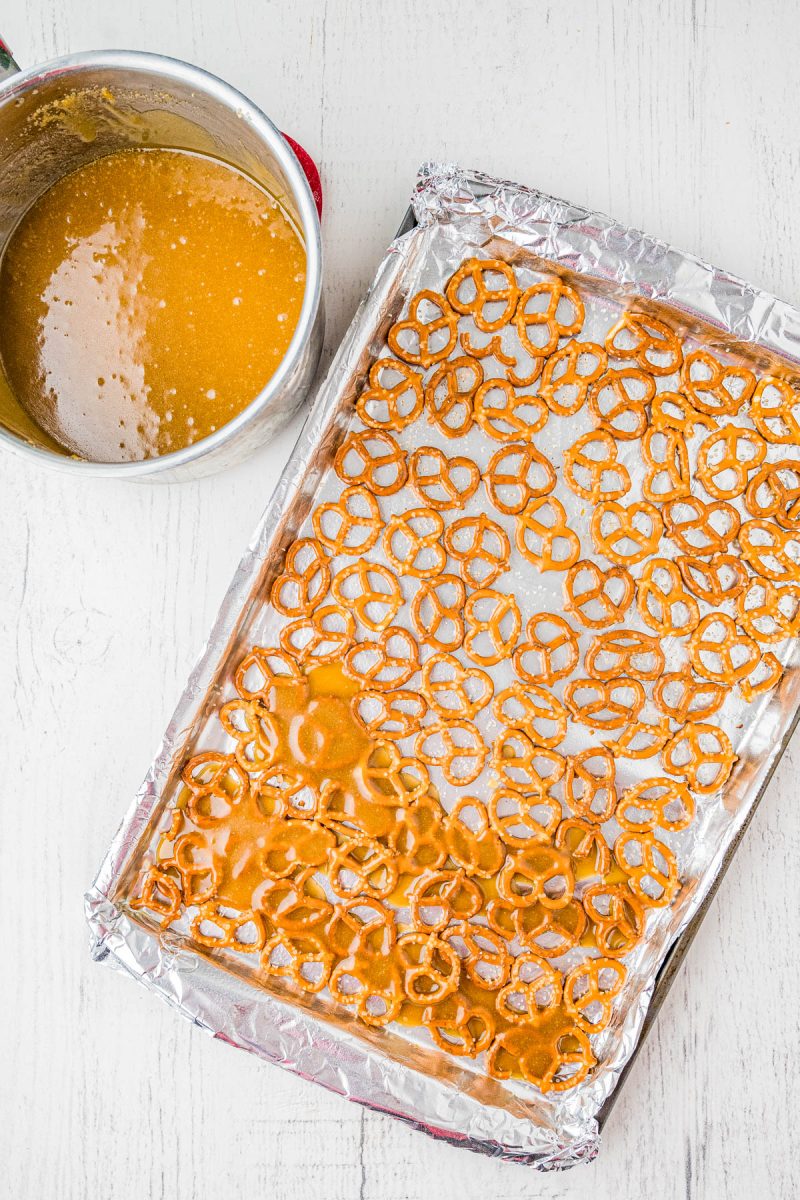 A baking sheet lined with pretzels is partially covered with caramel sauce; a saucepan with more caramel is nearby on a white surface.