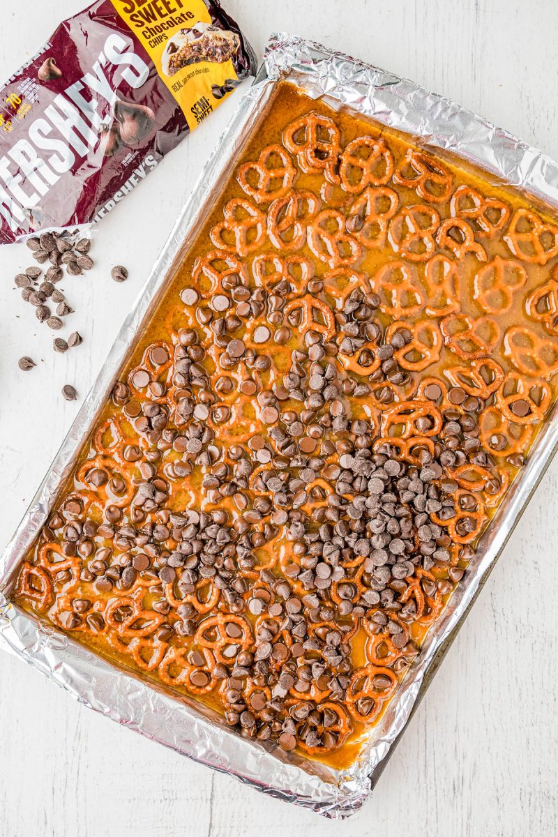 A baking sheet lined with foil holds a layer of pretzels, toffee, and scattered chocolate chips. A partially opened bag of Hershey’s milk chocolate chips and loose chips sit nearby.