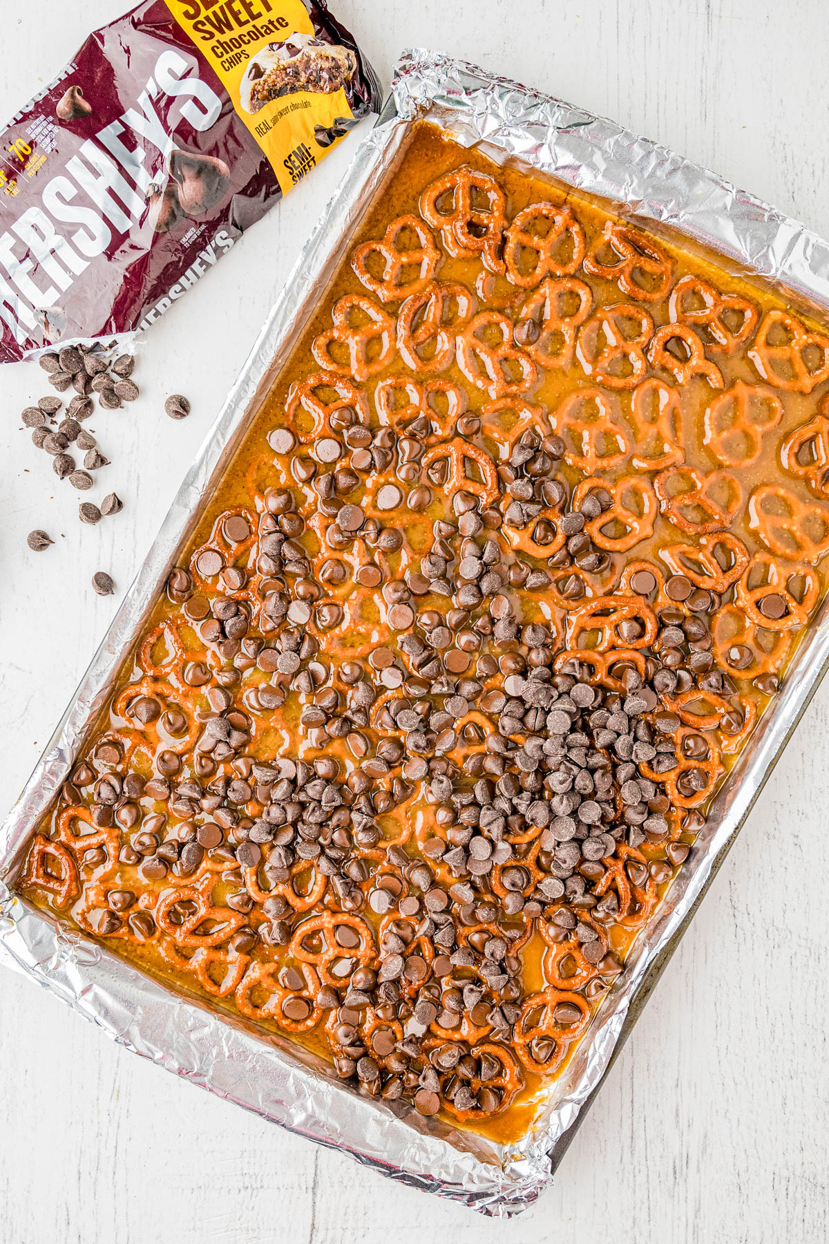 A baking sheet lined with foil holds a layer of pretzels, toffee, and scattered chocolate chips. A partially opened bag of Hersheyโs milk chocolate chips and loose chips sit nearby.