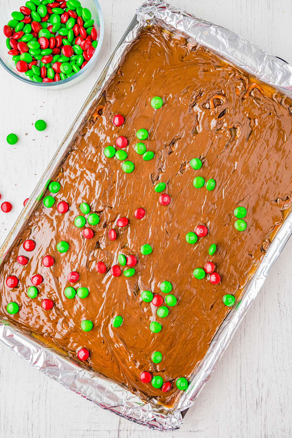 A baking sheet covered with chocolate toffee, topped with red and green M&Ms, sits on a white surface next to a bowl of additional M&Ms.