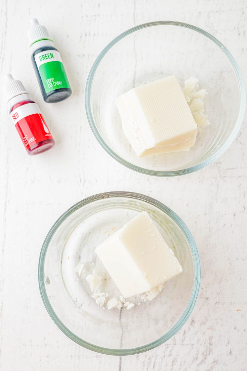 Two glass bowls each holding a block of white soap base, placed beside small bottles of red and green liquid soap coloring on a white surface.