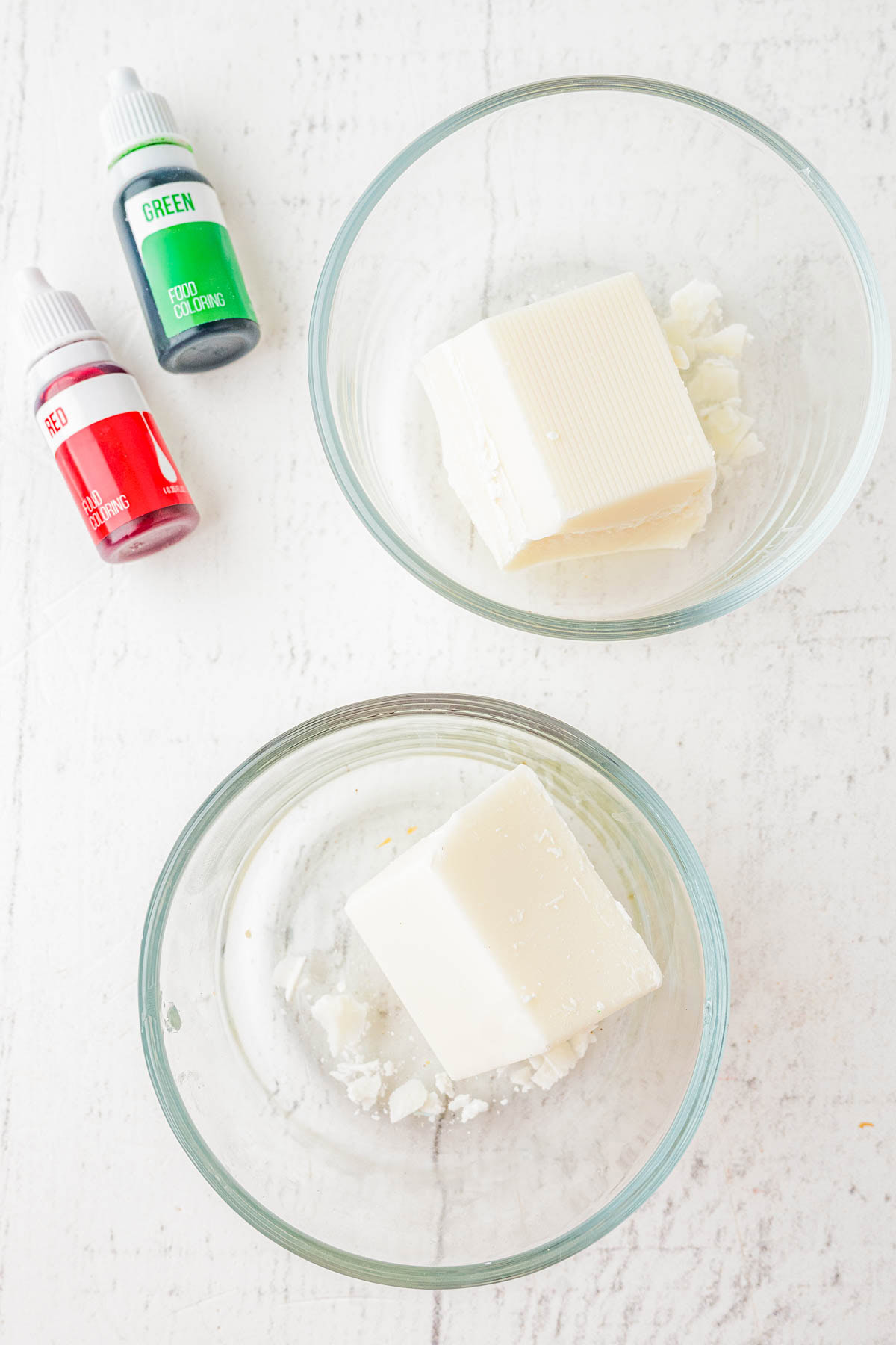 Two glass bowls each holding a block of white soap base, placed beside small bottles of red and green liquid soap coloring on a white surface.