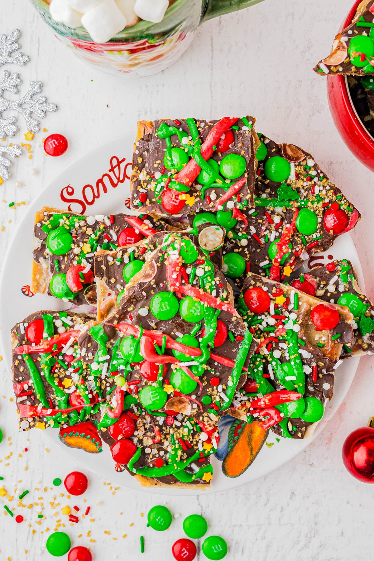 A plate of chocolate bark topped with red and green candies, sprinkles, and drizzled icing, displayed on a white surface with a "Santa" plate.