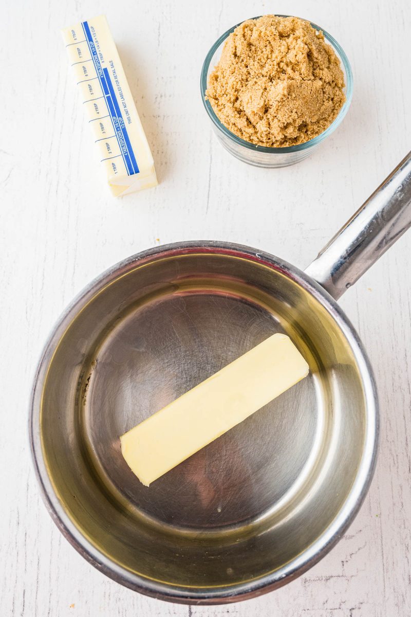 A stick of butter in a saucepan, with a bowl of brown sugar and another stick of butter on a white surface nearby.