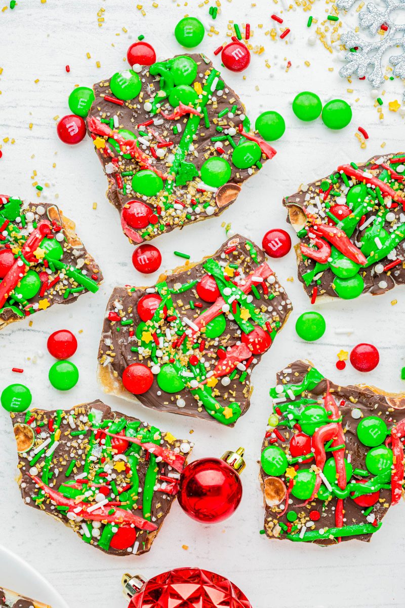 Pieces of chocolate bark topped with red and green candy, sprinkles, and nuts are arranged on a white surface with red and green Christmas ornaments nearby.