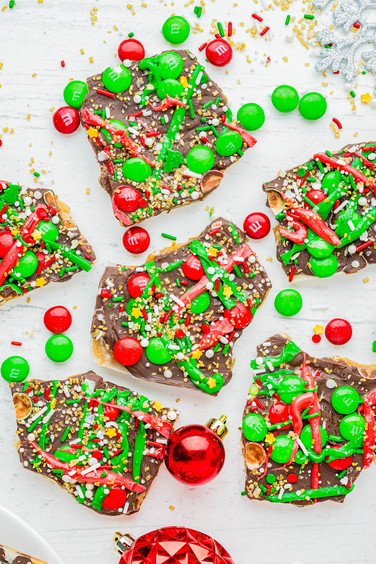 Pieces of chocolate bark topped with red and green candy, sprinkles, and nuts are arranged on a white surface with red and green Christmas ornaments nearby.