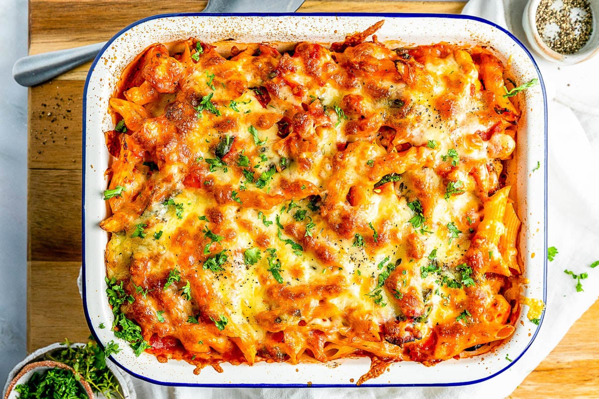 Baked pasta casserole topped with melted cheese and parsley, served in a white enamel dish on a wooden surface.