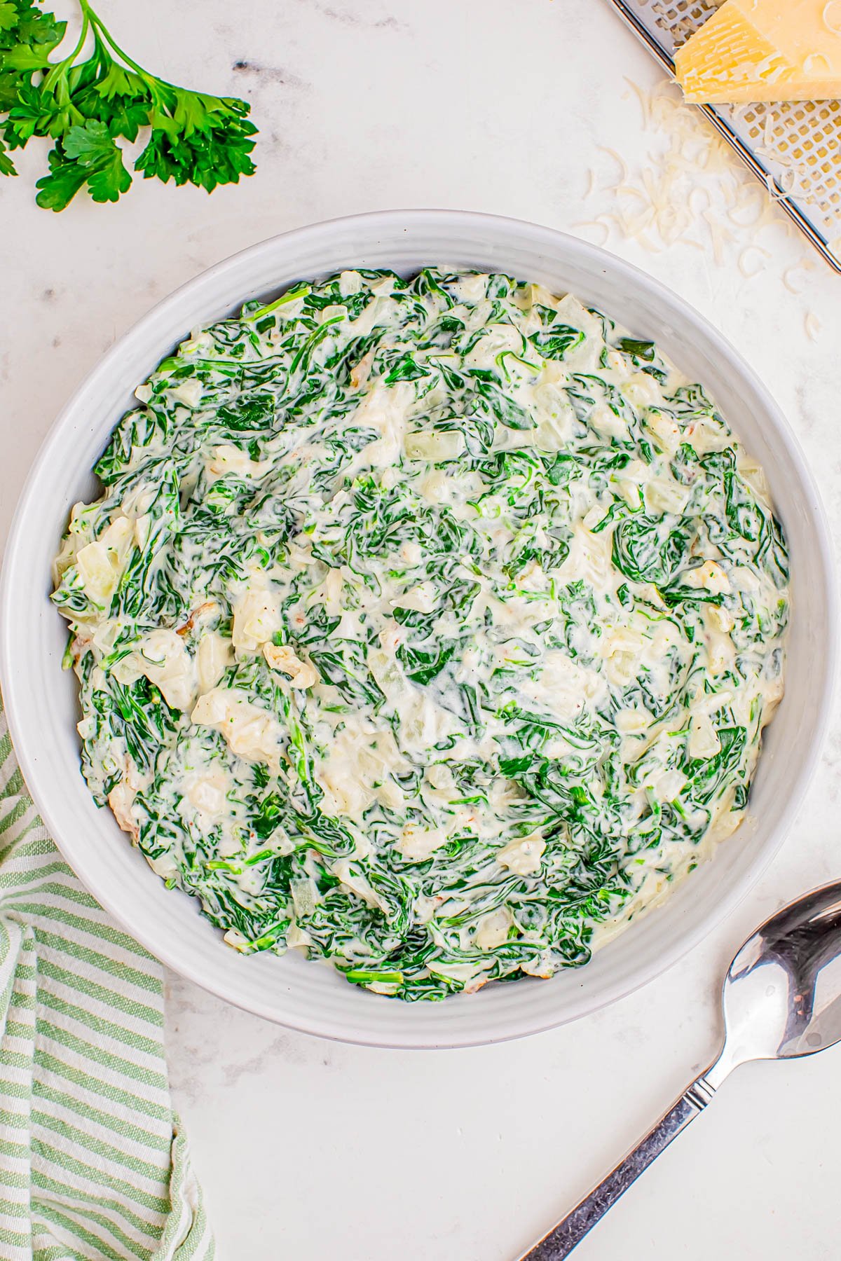 A white bowl filled with creamy spinach dip sits on a marble surface, next to a striped towel, a spoon, parsley, and a metal cheese grater.