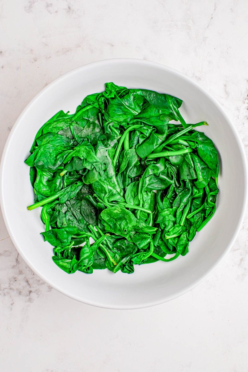 A white bowl filled with cooked spinach leaves on a light, textured surface.