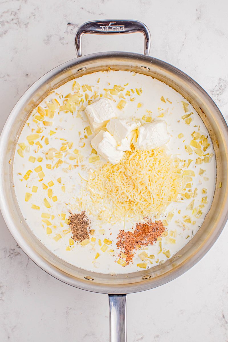 A stainless steel pan contains cream, chopped onions, shredded cheese, cream cheese, black pepper, and ground spices on a white marble surface.
