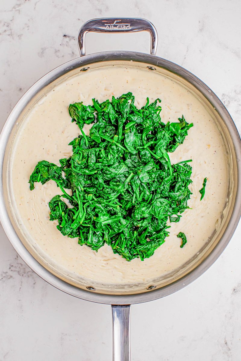 A stainless steel pan containing creamy white sauce with cooked spinach piled in the center, placed on a light marble surface.