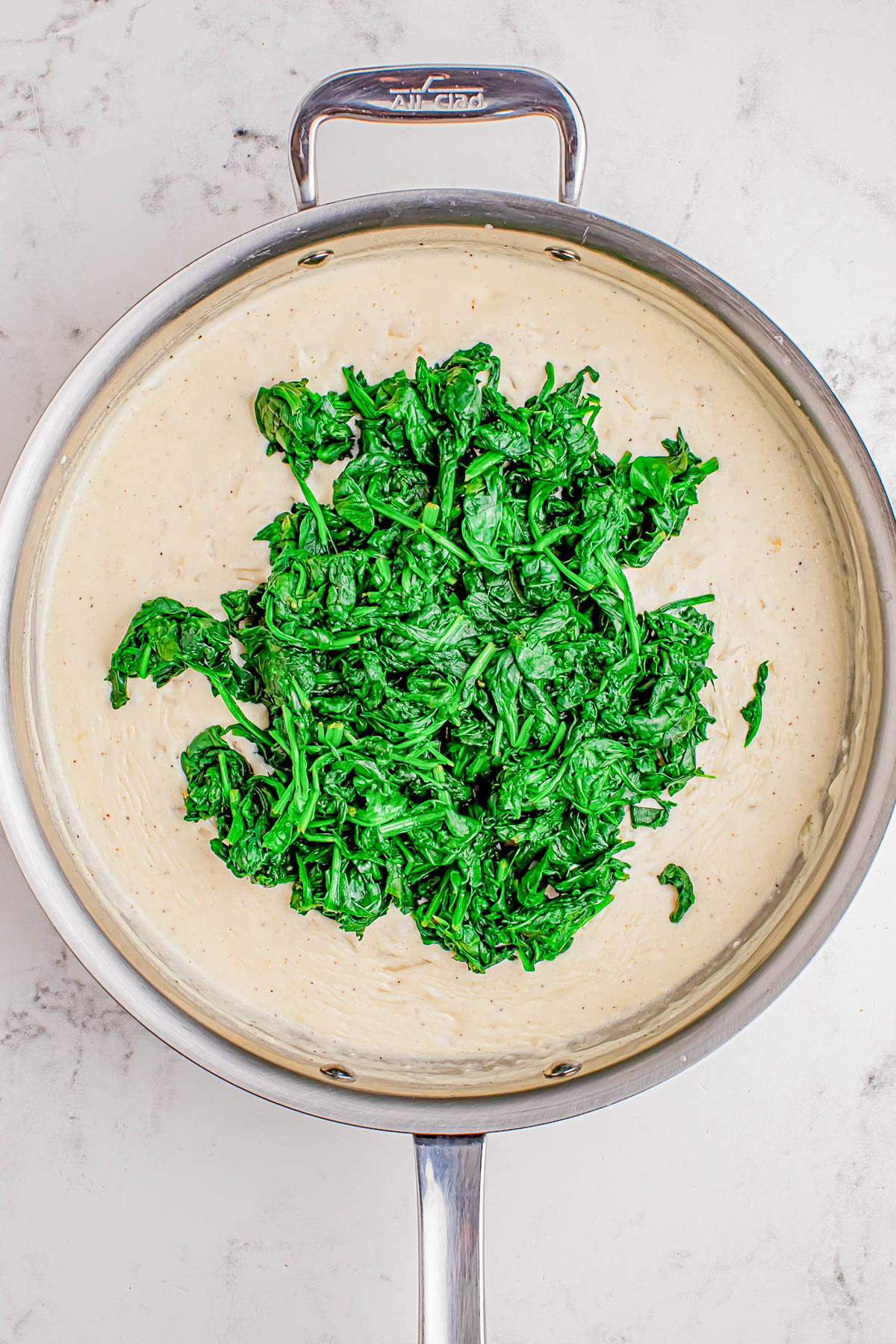 A stainless steel pan containing creamy white sauce with cooked spinach piled in the center, placed on a light marble surface.