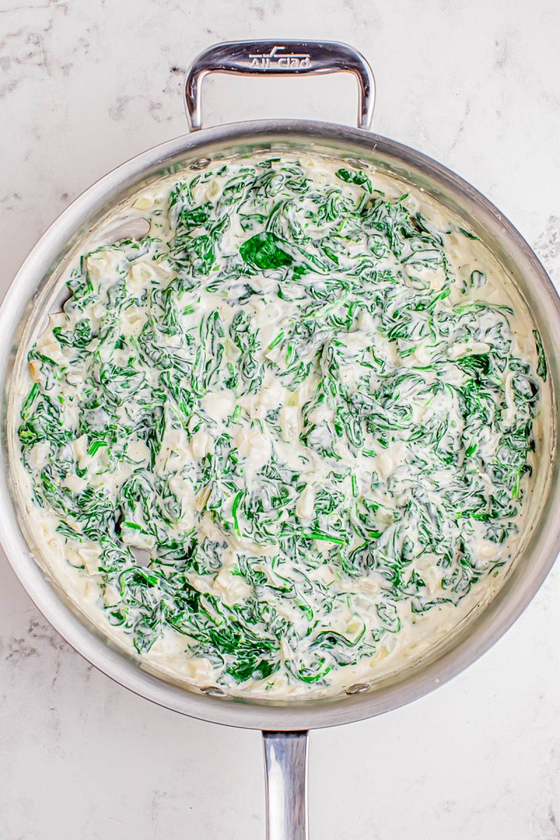 A stainless steel skillet filled with creamy spinach dip sits on a light marble surface.