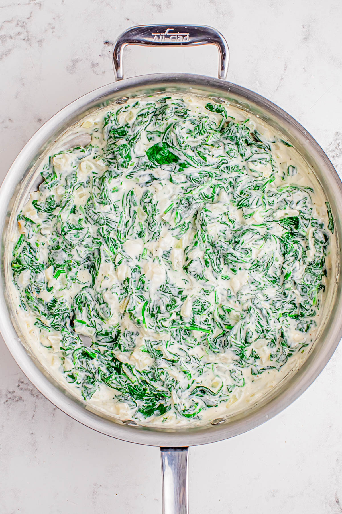 A stainless steel skillet filled with creamy spinach dip sits on a light marble surface.