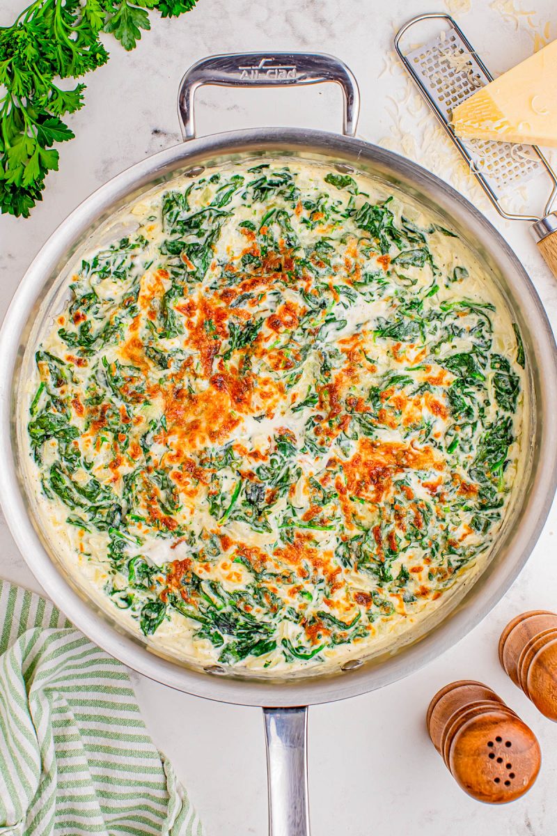 A skillet filled with creamy spinach and cheese gratin, topped with melted, lightly browned cheese. Grated cheese, parsley, salt and pepper shakers, and a striped towel are nearby.