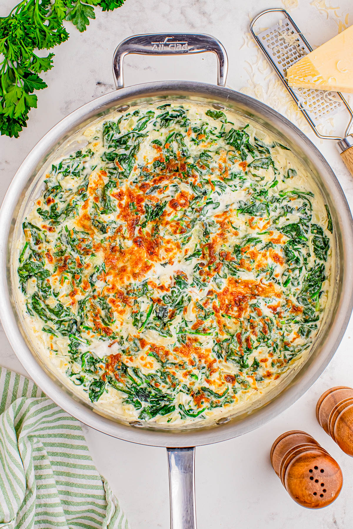 A skillet filled with creamy spinach and cheese gratin, topped with melted, lightly browned cheese. Grated cheese, parsley, salt and pepper shakers, and a striped towel are nearby.