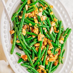 Oval white dish filled with green beans topped with toasted sliced almonds; a serving spoon rests in the dish.