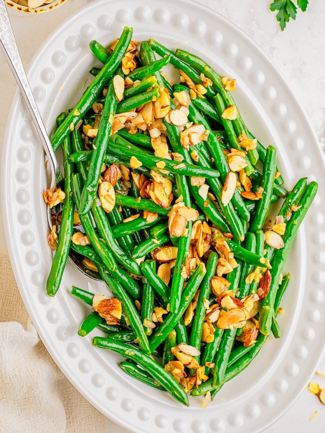 Oval white dish filled with green beans topped with toasted sliced almonds; a serving spoon rests in the dish.