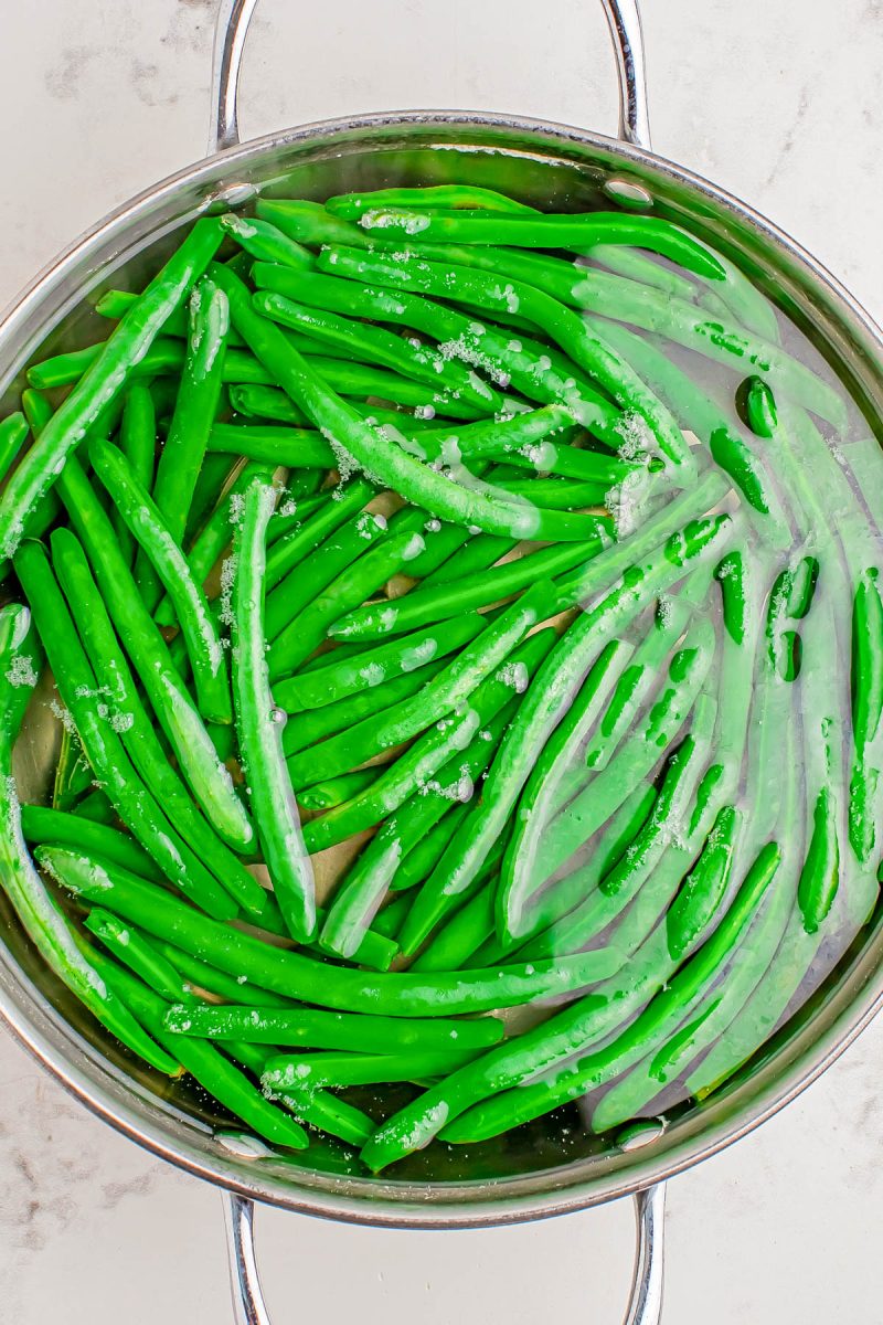 Green beans are submerged in water in a stainless steel pot with ice on the surface, suggesting they are being blanched or cooled.