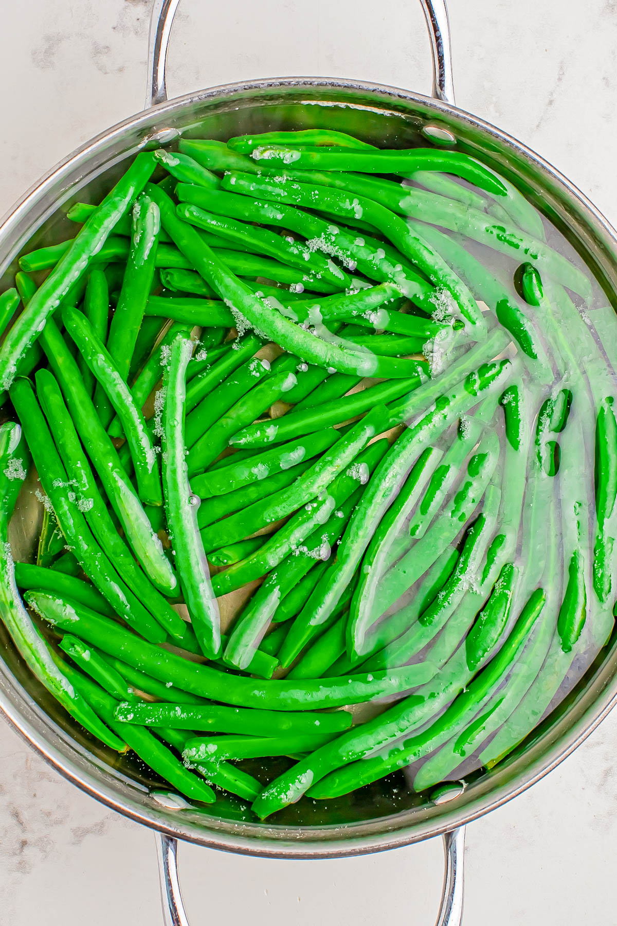 Green beans are submerged in water in a stainless steel pot with ice on the surface, suggesting they are being blanched or cooled.
