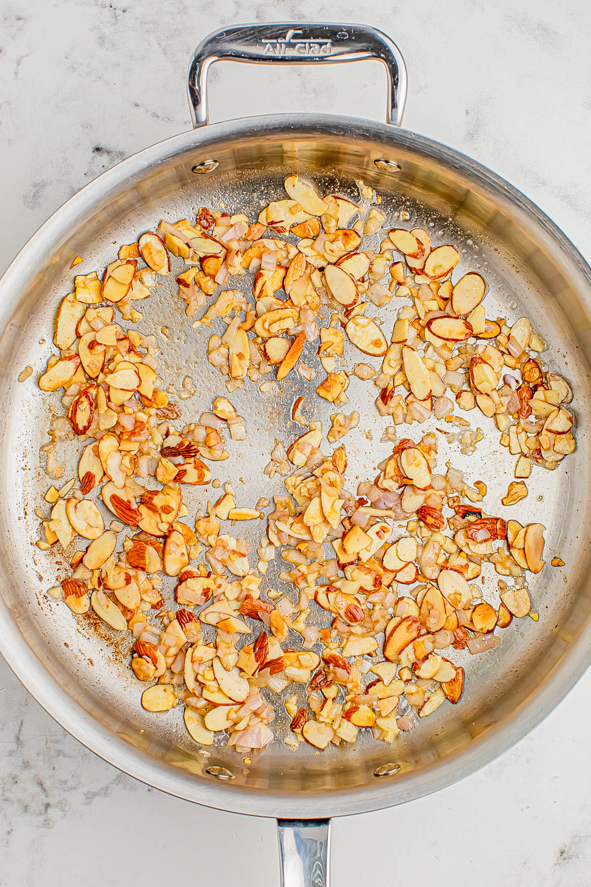 A stainless steel pan with sautรฉed sliced almonds and chopped onions on a light surface.