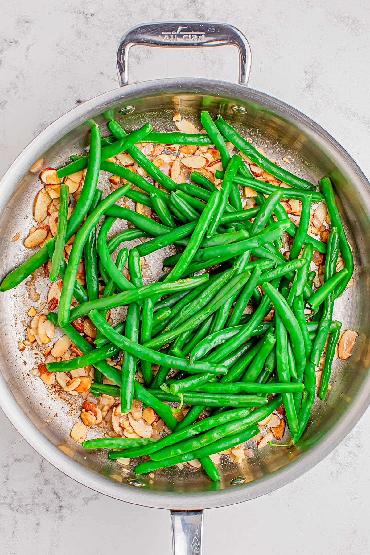 Green beans and sliced almonds are being sautรฉed in a stainless steel skillet on a marble countertop.