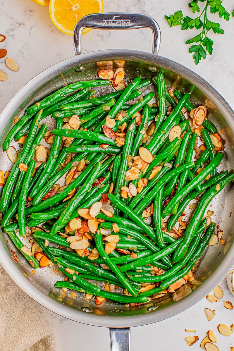 Sautéed green beans with sliced almonds in a stainless steel pan, garnished with herbs, and lemon slices in the background.