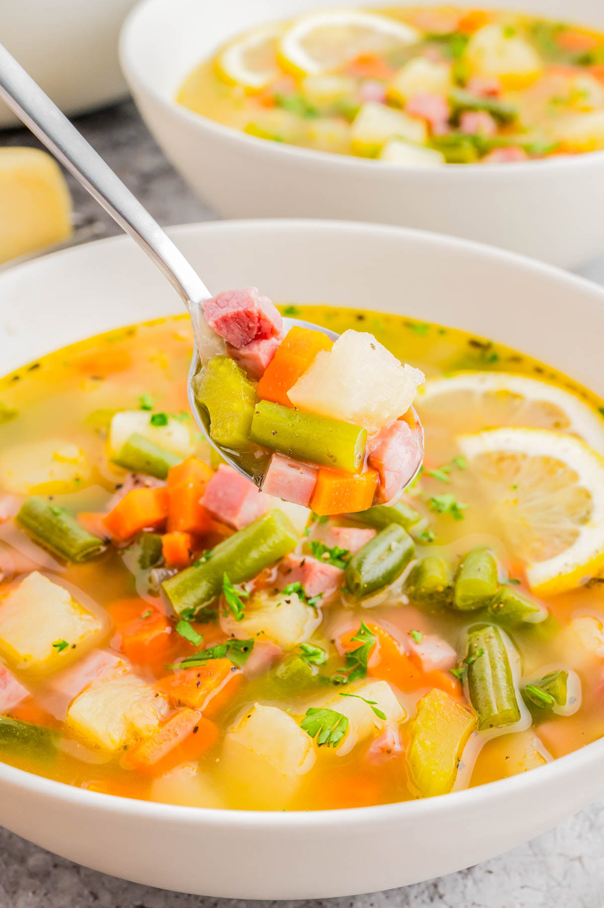 A bowl of vegetable soup with diced ham, carrots, green beans, potatoes, and lemon slices, with a spoonful lifted above the bowl.
