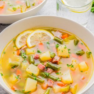 A bowl of vegetable soup with potatoes, carrots, green beans, diced ham, and lemon slices, garnished with herbs. A glass of water with a lemon slice sits nearby.
