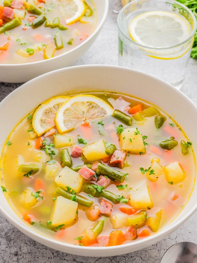 A bowl of vegetable soup with potatoes, carrots, green beans, diced ham, and lemon slices, garnished with herbs. A glass of water with a lemon slice sits nearby.