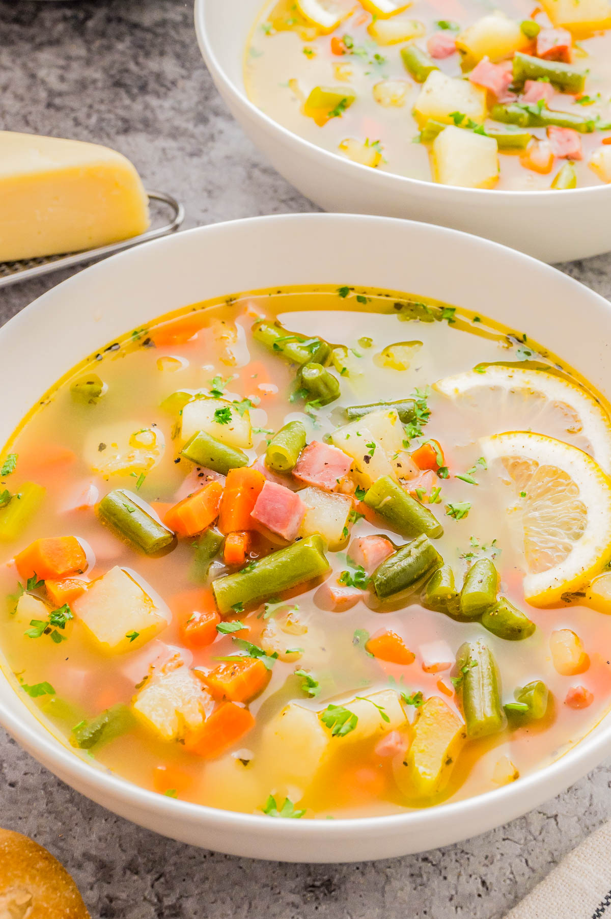 A bowl of vegetable soup with diced ham, green beans, carrots, potatoes, and lemon slices, garnished with chopped herbs.