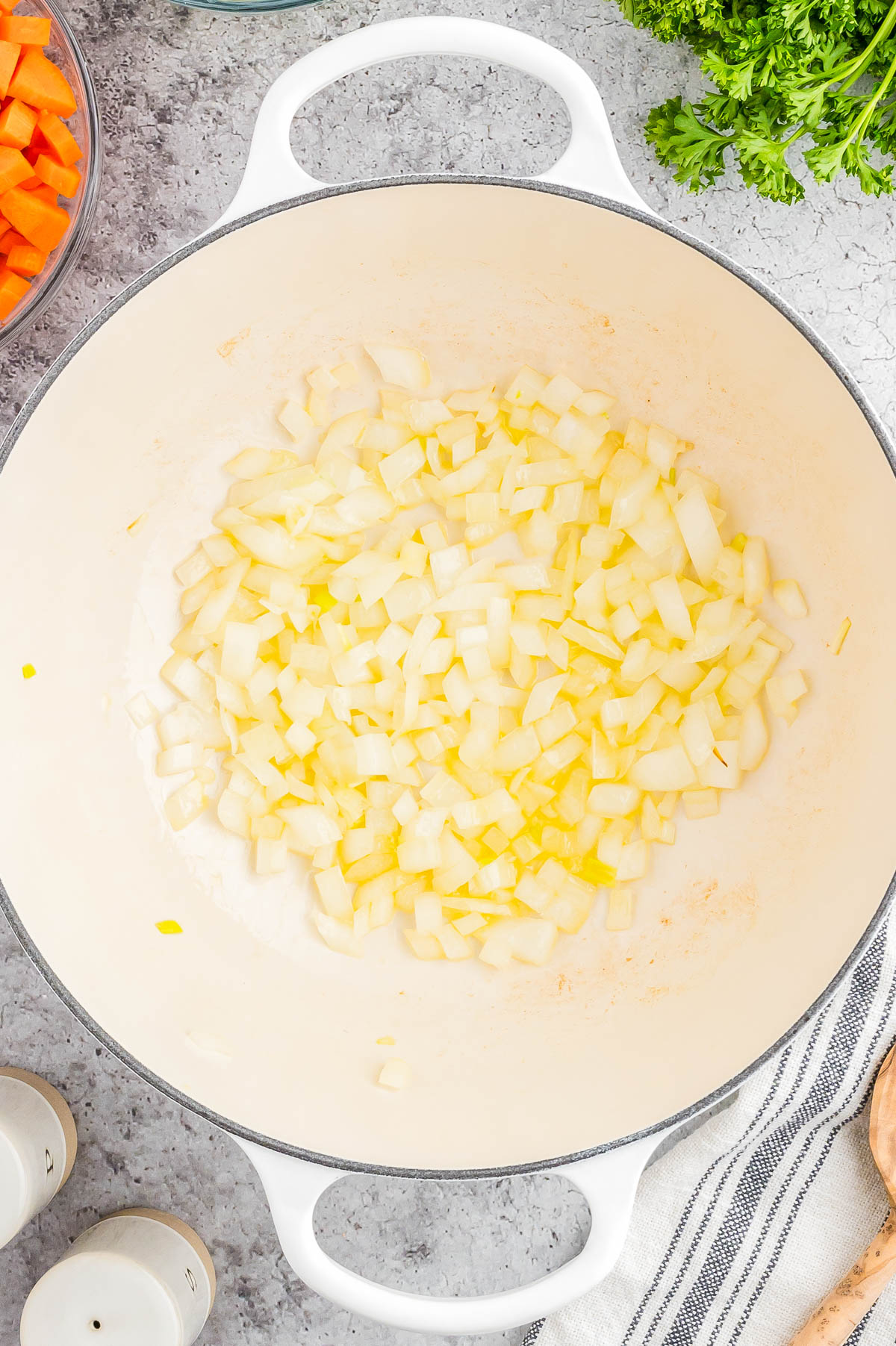 Chopped onions sautéing in a large white pot, surrounded by fresh vegetables and kitchen items on a countertop.