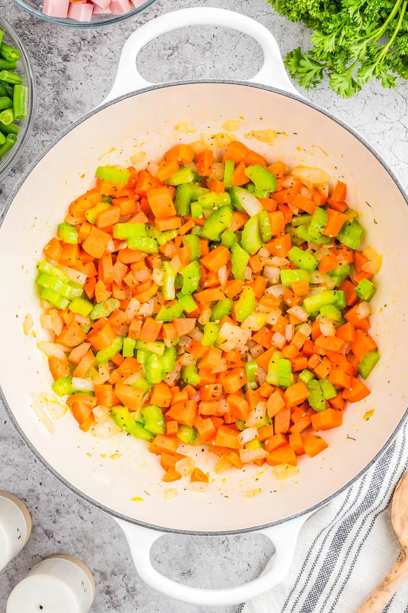Chopped carrots, celery, and onions are sautéing in a large white pot on a gray countertop, surrounded by various ingredients.