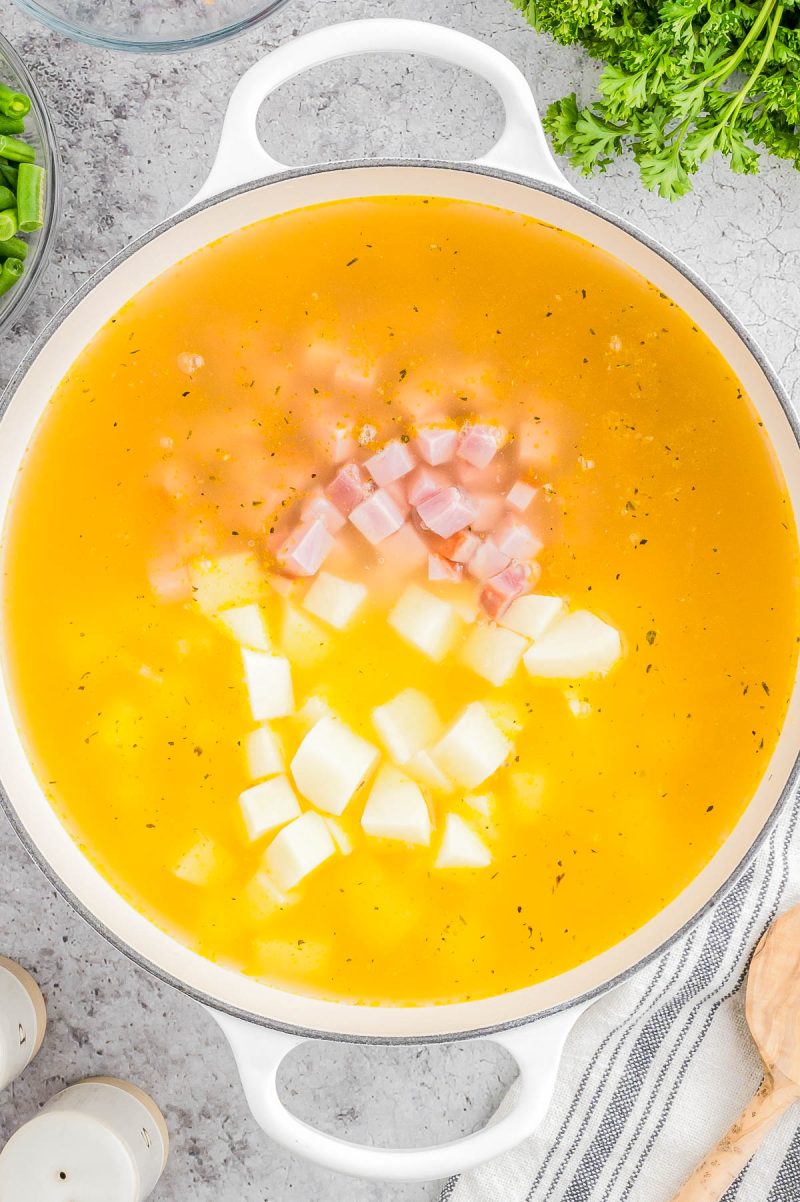 A pot of soup with diced ham and potatoes in a yellow broth, surrounded by parsley and kitchen items on a gray surface.
