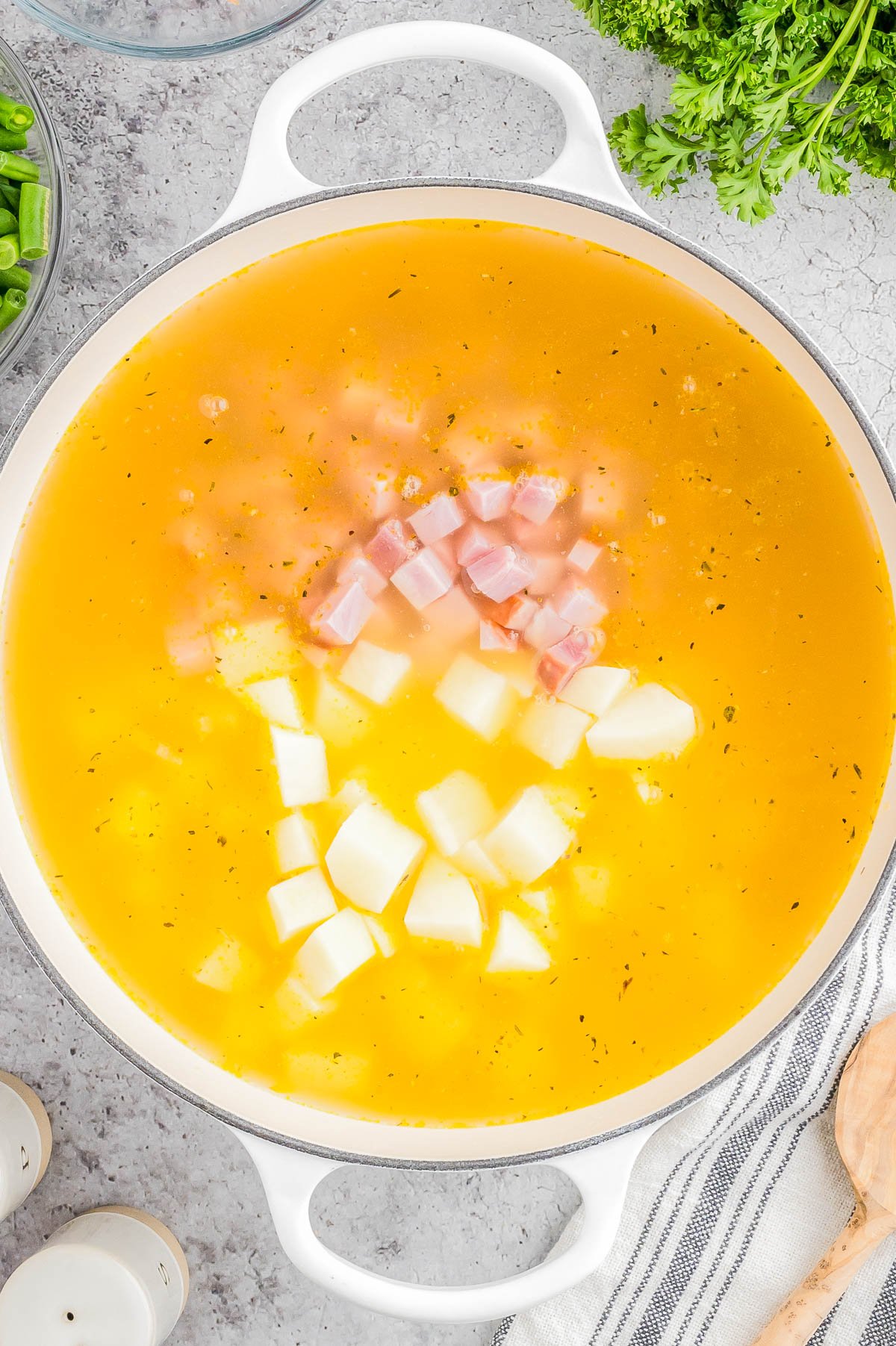 A pot of soup with diced ham and potatoes in a yellow broth, surrounded by parsley and kitchen items on a gray surface.